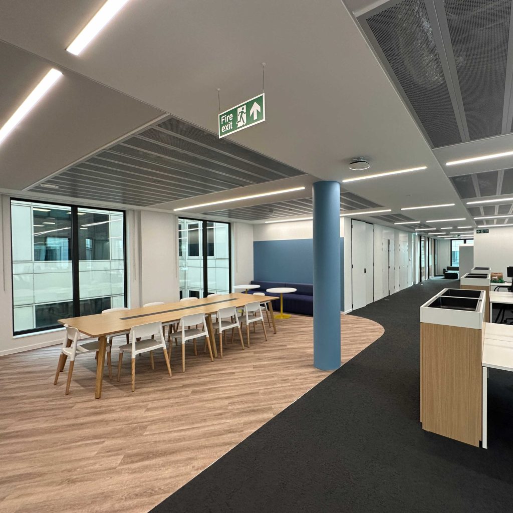 A modern office space after an office refurbishment, showing a long meeting table, a blue booth, and a fire exit sign.