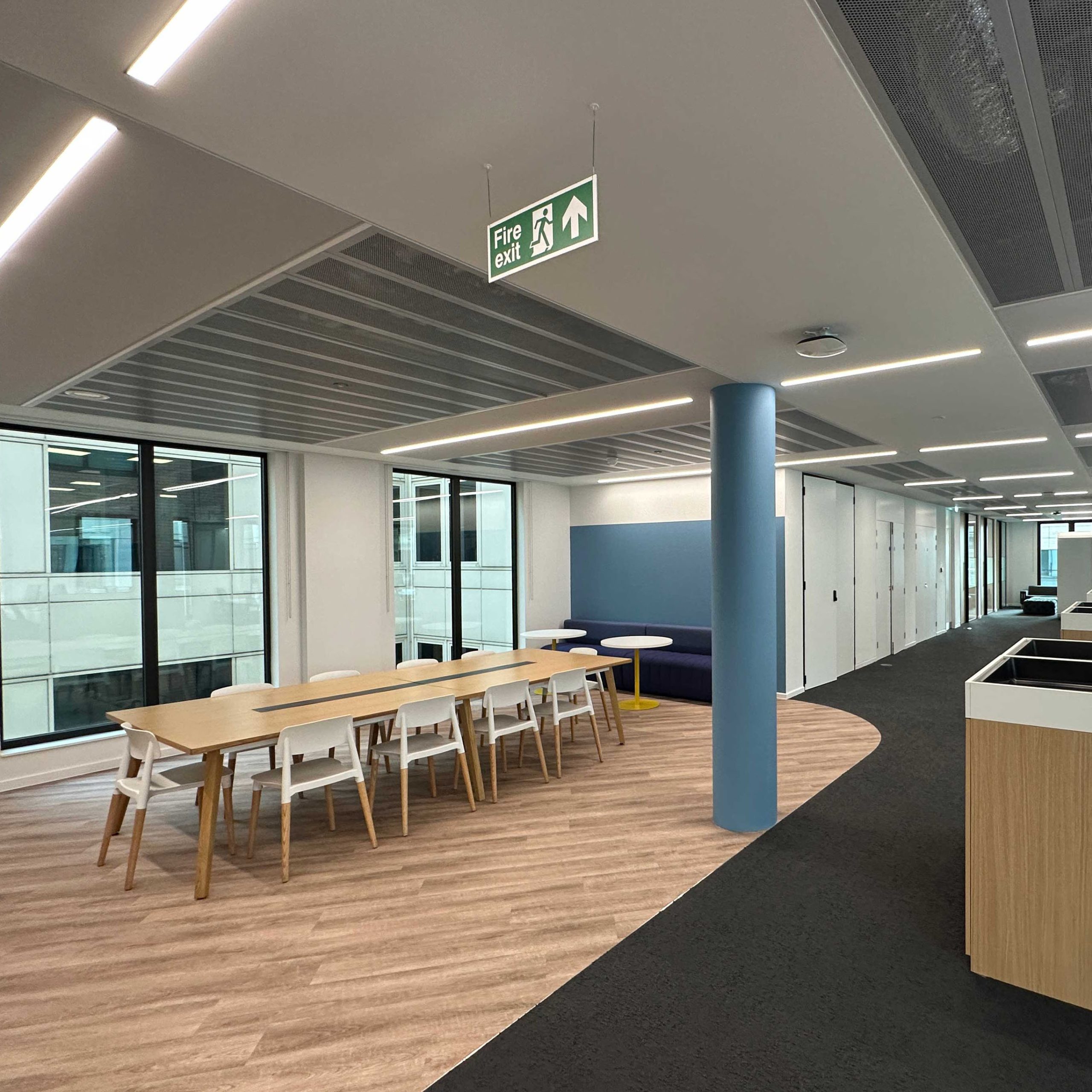 A modern office space after an office refurbishment, showing a long meeting table, a blue booth, and a fire exit sign.